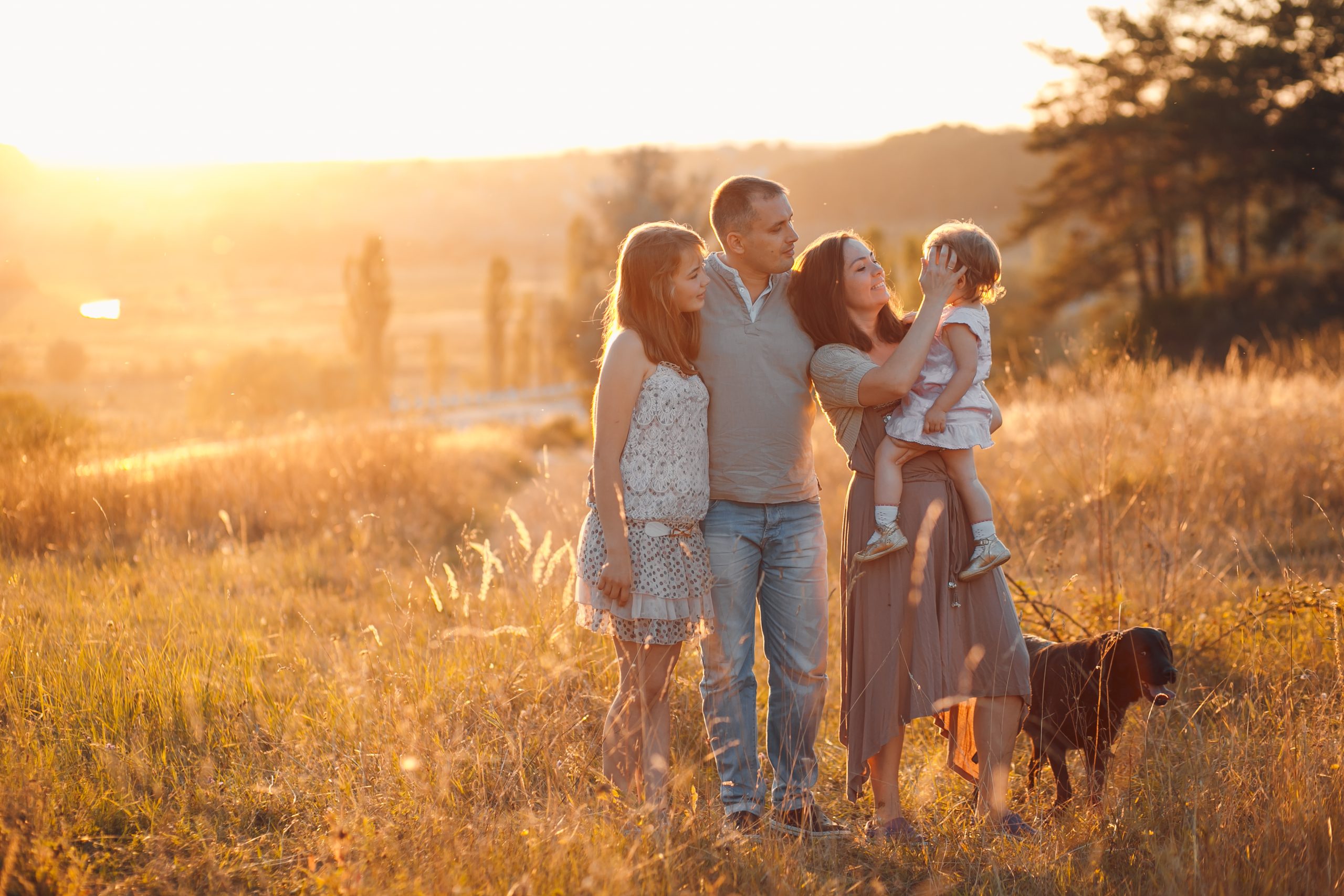 Family with two children and a dog standing together in a rural field at sunset